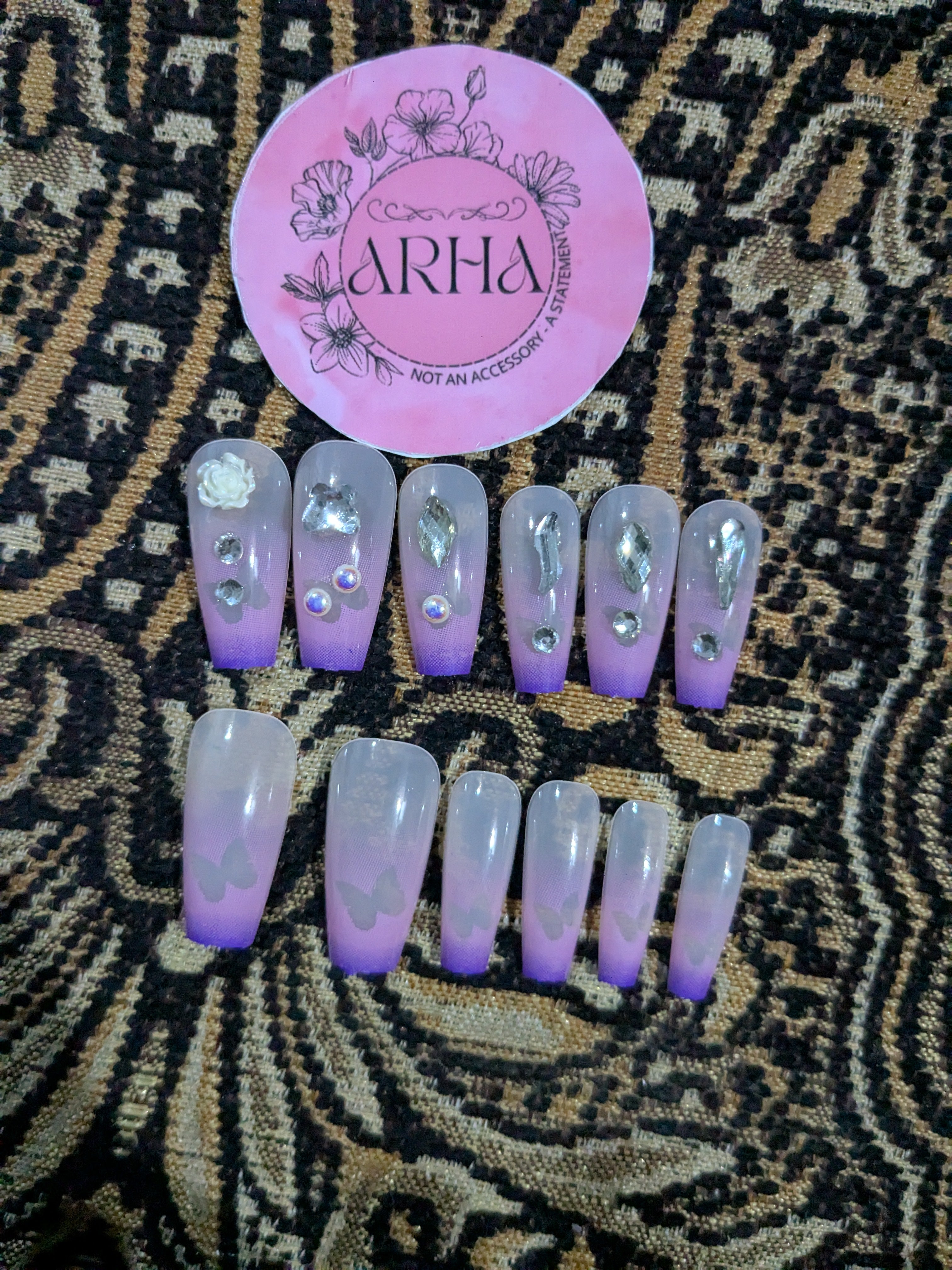 Set of purple and clear false nails with decorative elements on a patterned surface, featuring the brand 'ARHA'.